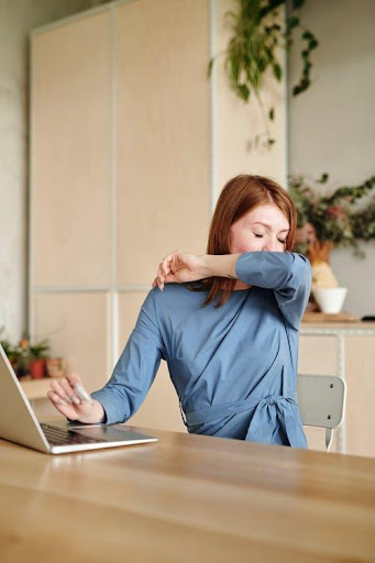 woman sitting at desk in front of laptop sneezing into elbow