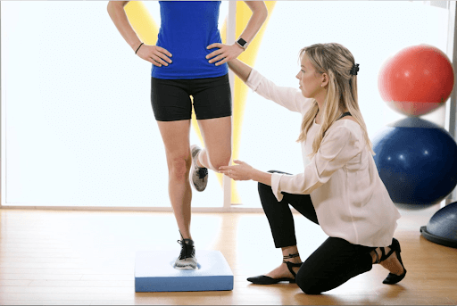 person balancing on one leg on blue foam pad with physiotherapist crouching beside and using her hands to cue balance