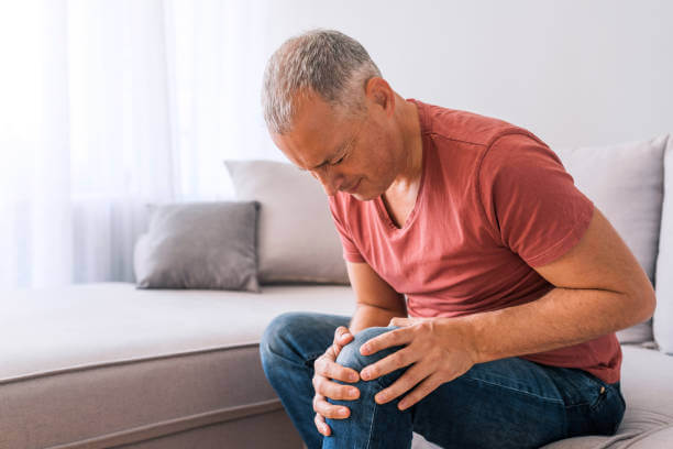 Photo of mature, elderly man sitting on a sofa in the living room at home, touching his knee
