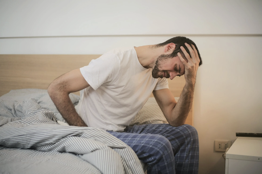 male sitting on edge of bed with hand on head looking uncomfortable